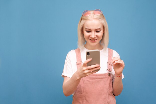 Glückliche junge dame in der niedlichen kleidung, die auf einem blauen hintergrund steht, ein smartphone benutzt und lächelt, sonnenbrille, ein weißes t-shirt und ein rosa kleid tragend