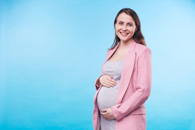 Glückliche junge elegante zukünftige Mutter im grauen lässigen Kleid und im rosa Trenchcoat, die Hände ihres Bauches halten, während sie isoliert stehen