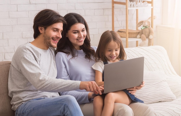 Glückliche junge Eltern mit kleiner Tochter, die zusammen Laptop benutzen, während sie zu Hause auf der Couch sitzen, im Internet surfen oder einen Film schauen.