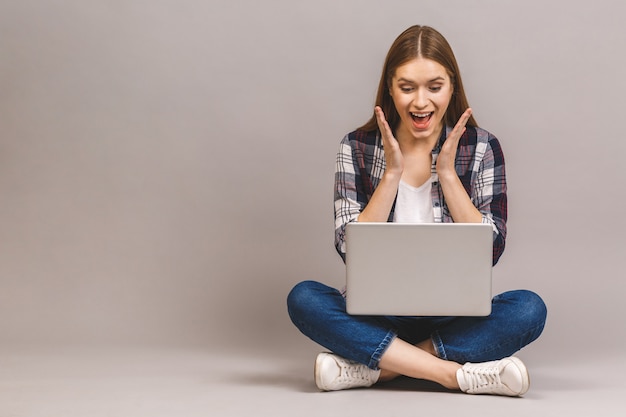 Glückliche junge erstaunte Frau, die auf dem Boden mit gekreuzten Beinen sitzt und Laptop benutzt