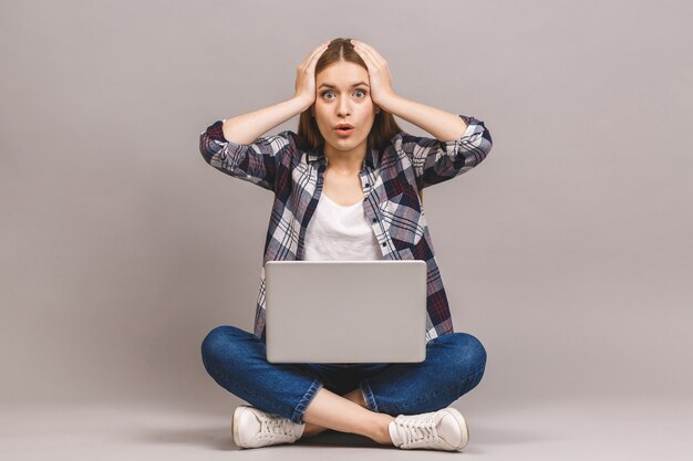 Glückliche junge erstaunte Frau, die auf dem Boden mit gekreuzten Beinen sitzt und Laptop benutzt