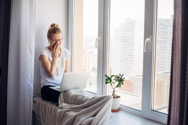 Glückliche junge erwachsene Frau Freiberufler mit Brille sitzt auf Fensterhintergrund Lachende Blondine arbeitet am Laptop Arbeitsplatz auf der Fensterbank Home-Office-Konzept