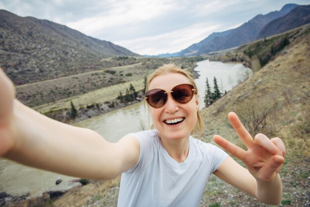 Glückliche junge erwachsene Frau mit Sonnenbrille und weißem T-Shirt macht ein Selfie und lacht in eine Smartphone-Kamera gegen die Bergflusslandschaft. Reisen, Bloggen, aktiver Lebensstil.
