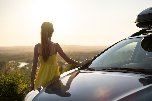 Glückliche junge Fahrerin im gelben Kleid, die sich auf ihr Auto stützt und einen warmen Sommertag genießt. Reise- und Urlaubskonzept.