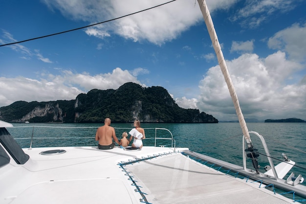 Glückliche junge Familie in weißer Kleidung - langhaarige blonde Frau, kleines Kind und kahler Mann, die zusammen auf Yacht sitzen und das blaue Meer betrachten. Phuket. Thailand