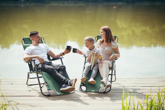 Glückliche junge Familie mit Kleinkindkindern, die draußen auf klappbaren Campingstühlen über dem See auf einem Holzsteg sitzen. Verbringen Sie gemeinsame Freizeit im Camp Ponton mit Kindern in der Natur. Ferienwald