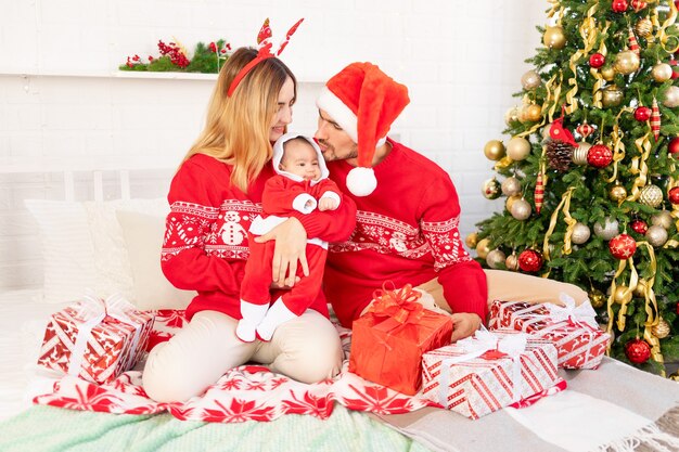 Glückliche junge Familienmutter, Papa und Baby in roten Pullovern unter dem Weihnachtsbaum auf dem Bett zu Hause feiern Neujahr oder Weihnachten, indem sie sich küssen und umarmen
