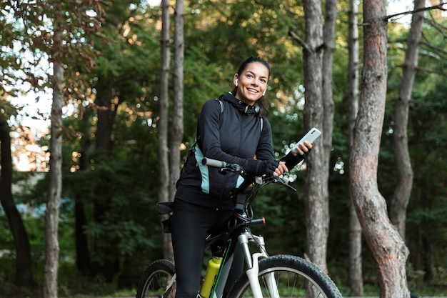 Glückliche junge Fitnessfrau, die auf einem Fahrrad am Park reitet, Musik mit Kopfhörern hörend, Handy hält