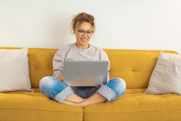 Glückliche junge Frau, die am Laptop sitzt auf Sofa arbeitet. Frau, die von zu Hause aus arbeitet. Junge Frau mit Brille lächelt während der Arbeit am Laptop mit gekreuzten Beinen auf dem Sofa zu Hause.