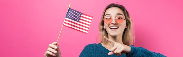 Glückliche junge Frau, die amerikanische Flagge gegen einen rosa Studiohintergrund hält