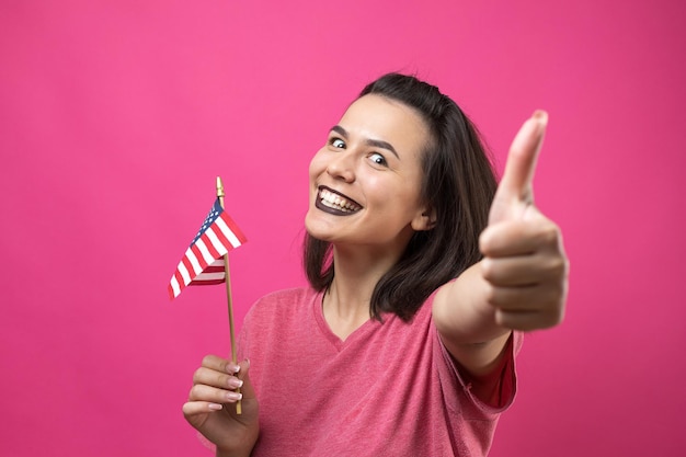 Glückliche junge Frau, die amerikanische Flagge gegen einen rosa Studiohintergrund hält