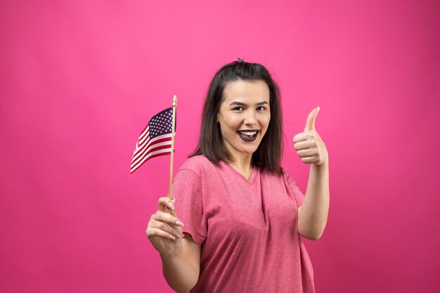 Glückliche junge Frau, die amerikanische Flagge gegen einen rosafarbenen Hintergrund des Studios hält