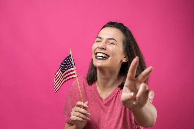 Glückliche junge Frau, die amerikanische Flagge gegen einen rosafarbenen Hintergrund des Studios hält