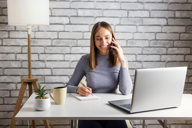 Glückliche junge Frau, die an ihrem Computer in einem weißen Schreibtisch und einer grauen Ziegelwand telefoniert