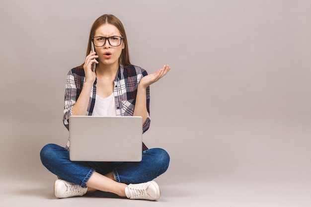 Glückliche junge Frau, die auf dem Boden mit gekreuzten Beinen sitzt und Laptop verwendet. Telefon benutzen.