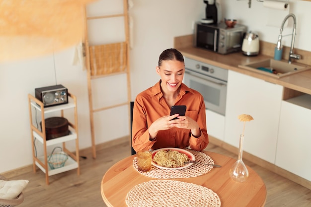 Glückliche junge Frau, die beim Mittagessen am Tisch im freien Küchenraum oben sitzt und ihr Mobiltelefon benutzt