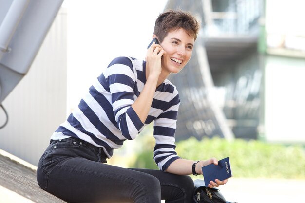 Glückliche junge Frau, die draußen mit Handy und Reisepass sitzt