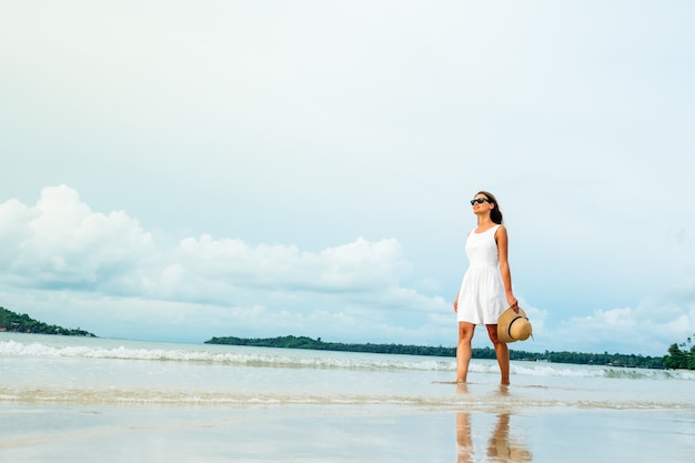 Glückliche junge Frau, die ein schönes weißes Kleid trägt, das am Strand spazieren geht