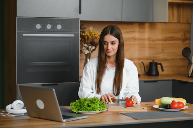 Glückliche junge Frau, die einen Salat in der Küche macht und Gemüse hackt und auf einen Laptop-Computer schaut