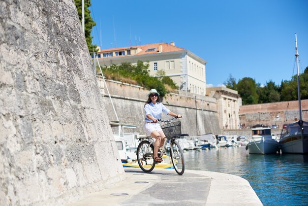 Glückliche junge Frau, die Fahrrad auf Steinweg entlang des Meeres durch hohe Steinmauer am hellen sonnigen Sommertag reitet. Sightseeing, Tourismus, Sport und aktives Lifestyle-Konzept.