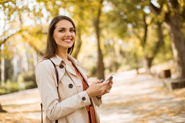 Glückliche junge Frau, die Handy im Herbstpark verwendet