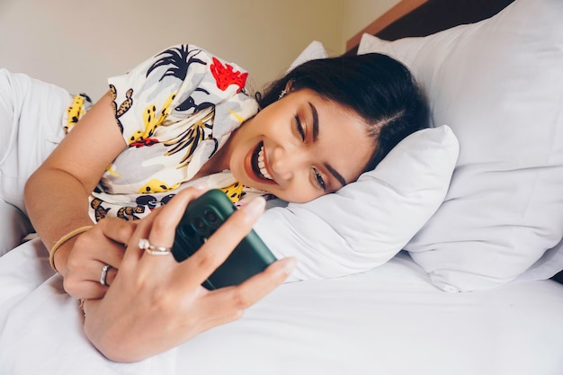 Glückliche junge Frau, die ihr Telefon überprüft, während sie im Bett liegt