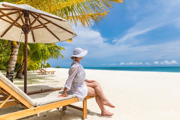 Glückliche junge Frau, die im weißen Kleid auf einem Liegestuhl am tropischen Strand sitzt. Das Leben genießen, unbeschwert