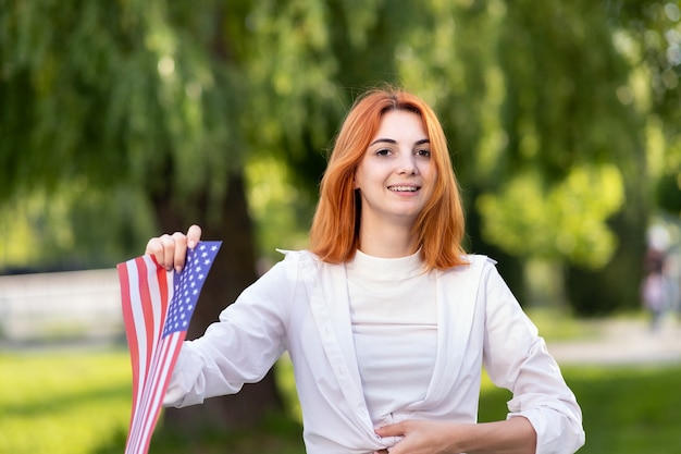 Glückliche junge Frau, die mit der Nationalflagge der USA aufwirft, die sie in ihrer ausgestreckten Hand hält, die draußen im Sommerpark steht. Hübsches Mädchen, das den Unabhängigkeitstag der Vereinigten Staaten feiert.