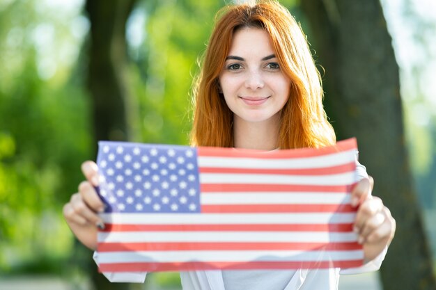 Glückliche junge Frau, die mit USA-Nationalflagge aufwirft, die es in ihren ausgestreckten Händen hält, die draußen im Sommerpark stehen.