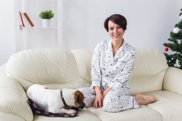 Glückliche junge Frau, die Pyjama mit reizendem Hund im Wohnzimmer mit Weihnachtsbaum trägt. Ferien