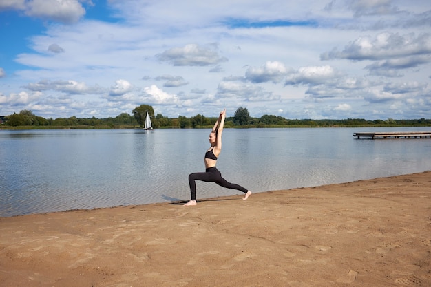 Glückliche junge Frau, die schwarze Sportkleidung trägt, die Yoga auf dem Sand nahe See im Freien praktiziert.