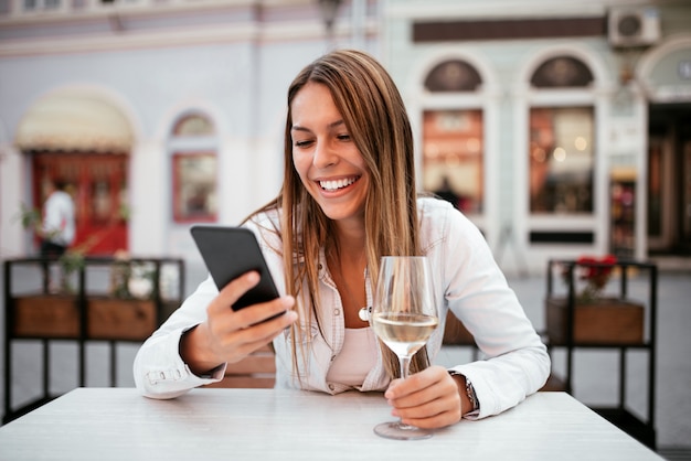 Glückliche junge Frau, die Smartphone verwendet und draußen ein Glas Weißwein im Restaurant isst.