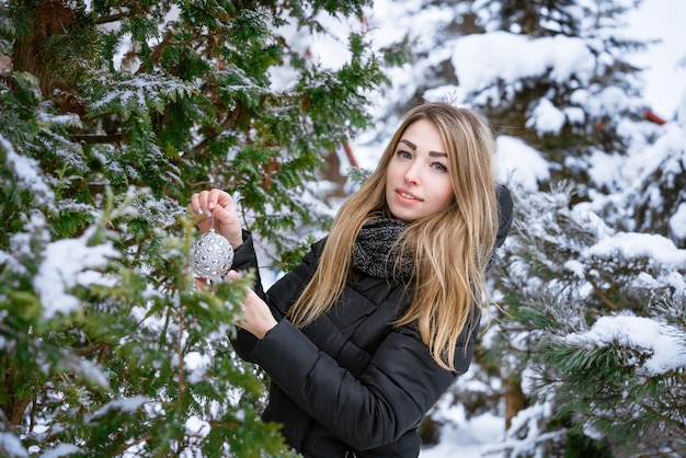 Glückliche junge Frau, die Tannenzweige mit moderner Christbaumkugel schmückt, die den Weihnachtsbaum im Schnee schmückt ...