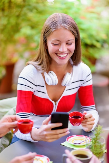 Glückliche junge Frau, die vor Freude lächelt, während sie ihr Smartphone benutzt, während sie sich an einem Tisch entspannt und mit Freunden in einem Restaurant im Freien eine Tasse Kaffee trinkt?