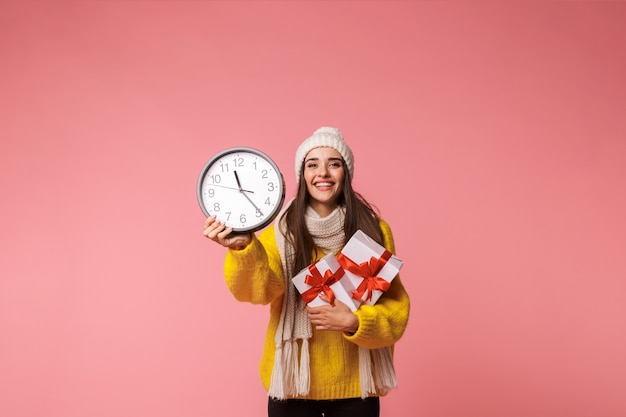 Glückliche junge Frau, die warmen Hut und Schal trägt, die lokal über Rosa stehen, Geschenkboxen halten, Uhr zeigend