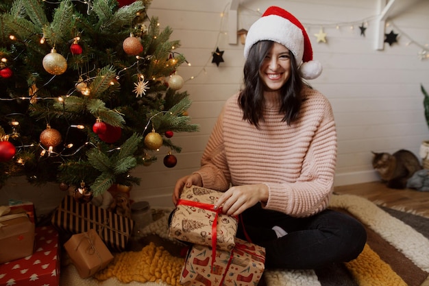 Glückliche junge Frau, die Weihnachtsgeschenkbox öffnet und unter Weihnachtsbaum mit Lichtern im modernen Zimmer lächelt Stilvolles Mädchen in Weihnachtsmütze, das Weihnachtsgeschenke hält Frohe Feiertage