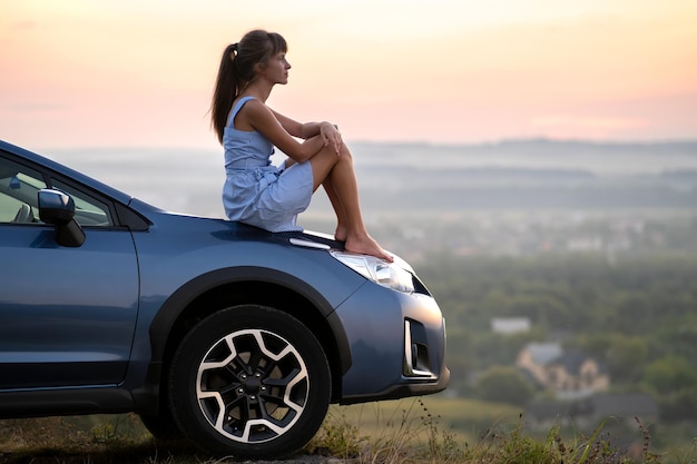 Glückliche junge Frau im blauen Kleid, die auf ihrer Fahrzeughaube sitzt und den Sonnenuntergang der Sommernatur betrachtet. Reise- und Urlaubskonzept.