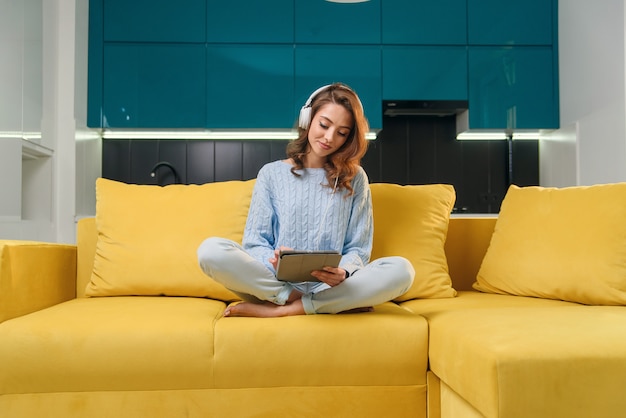 Glückliche junge Frau im blauen lässigen Pullover sitzt auf dem bequemen Sofa und verwendet Tablet-PC, um Musik in der zeitgenössischen Küche zu hören.