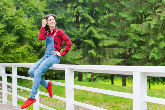 Glückliche junge Frau in blauem Jeansoverall und rotem Hemd sitzt auf einem Holzzaun