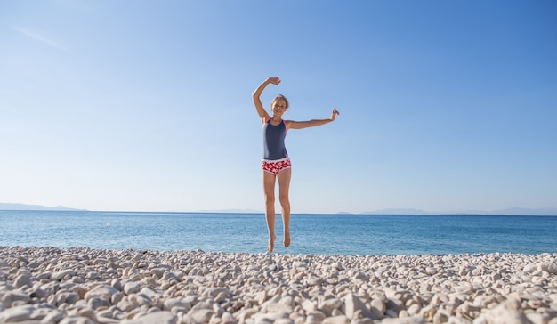 Glückliche junge Frau in der zufälligen Art, die am Steinstrand springt. Entspannen, Spaß haben und Urlaub am paradiesischen Strand mit blauem Himmel und Meer genießen. Mädchen in den Sommerferien.