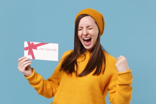 Glückliche junge Frau in gelber Strickmütze posiert isoliert auf blauem Hintergrund Studioportrait. Menschen aufrichtige Emotionen Lifestyle-Konzept. Mock-up-Kopienbereich. Geschenkgutschein mit Gewinnergeste halten.