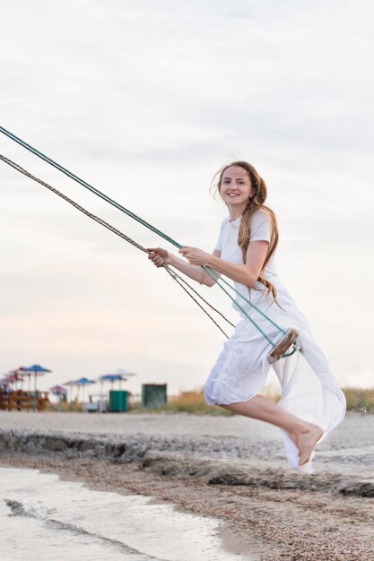 Glückliche junge Frau in weißem Kleid reitet auf einer Seilschaukel am Strand Vertikaler Rahmen