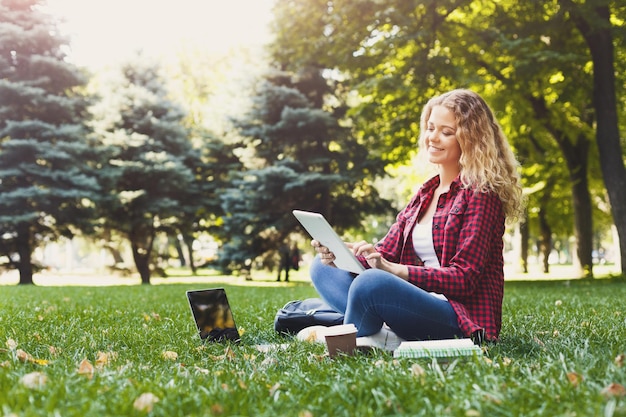 Glückliche junge Frau mit digitalem Tablet und Laptop im Freien. Studentin bereitet sich mit Computer, Büchern und Kaffee im Park auf Prüfungen vor. Bildung und Eintritt in das Universitätskonzept, Kopierraum.