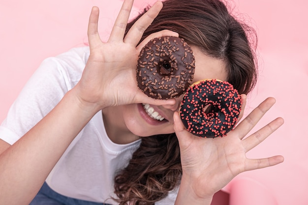 Glückliche junge Frau mit Donuts in ihren Händen lächelt, hält rosa Donuts in der Nähe ihrer Augen ihre Hände, rosa Hintergrund.