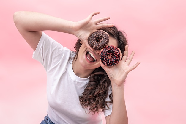 Glückliche junge Frau mit Donuts in ihren Händen lächelt, hält rosa Donuts in der Nähe ihrer Augen ihre Hände, rosa Hintergrund.