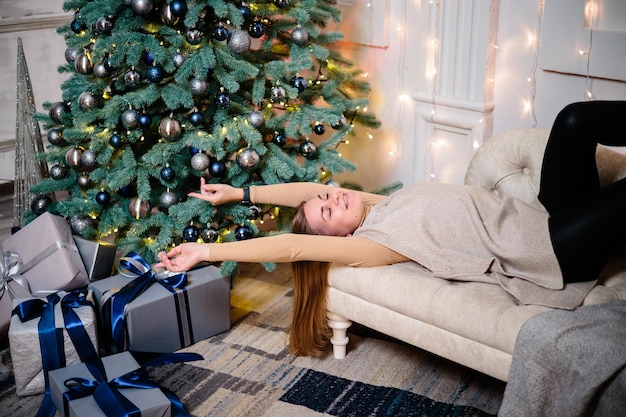 Glückliche junge Frau mit einem Geschenk, das nahe dem Weihnachtsbaum liegt Weiße silberne Farbe auf Sofahintergrund Frohe Weihnachten und neues Jahr Atmosphärische Momente