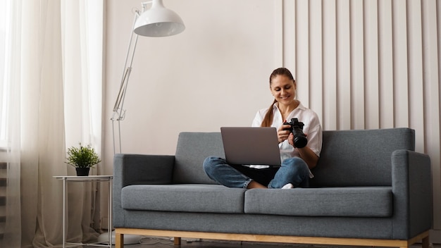 Glückliche junge frau mit fotokamera unter verwendung des laptops zu hause. der fotograf schaut sich die aufgenommenen bilder an und setzt sich auf die couch.