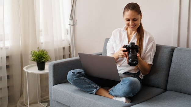 Glückliche junge Frau mit Fotokamera unter Verwendung des Laptops zu Hause. Der Fotograf schaut sich die aufgenommenen Bilder an und setzt sich auf die Couch.