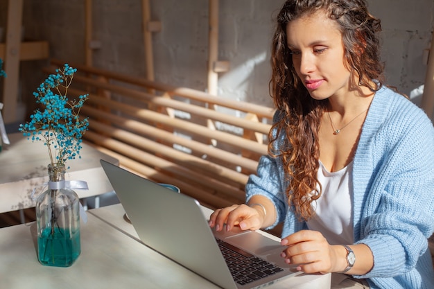 Glückliche junge Frau mit langen Haaren in blau gestricktem warmem Pullover, der nahe am Fenster sitzt und ferngesteuert zu Hause am Computer arbeitet. Wohnraum mit Fensterblick. Elegante Geschäftsfrau lächelt.