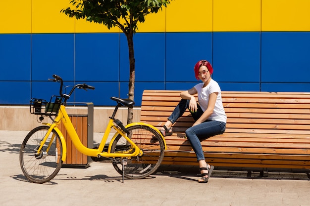 Glückliche junge Frau mit rosa Haaren geht im Sommer auf einem Fahrrad durch die Stadt.